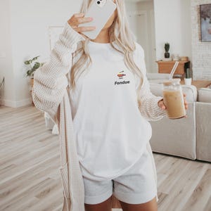 Puede incluir: Una camiseta blanca con un gráfico de una olla de fondue y la palabra "Fondue" impresa en la parte delantera. La persona lleva pantalones cortos grises y una chaqueta de punto crema.
