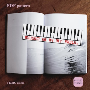 May include: A cross-stitch pattern featuring a piano keyboard design with the text "MUSIC IS IN MY SOUL" in red. The pattern is displayed on an open book with a grayscale wave image in the background. The words "PDF pattern" are at the top.