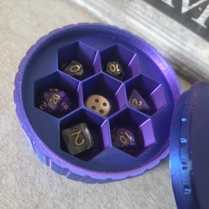 Könnte beinhalten: Eine runde, lila Würfelbox mit Wabenstruktur im Inneren, die mehrere vielseitige Würfel enthält. Die Würfel sind in verschiedenen Farben gehalten, darunter Schwarz, Lila und Gold, mit weißen oder goldenen Zahlen. Die Box ist geöffnet und zeigt die Würfel.