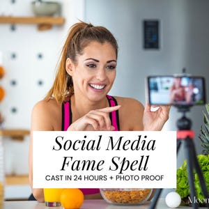 Op de afbeelding: Een vrouw in een roze en zwarte top glimlacht terwijl ze een smartphone op een statief vasthoudt. De telefoon toont een video. Tekst op de afbeelding luidt "Social Media Fame Spell" en "Cast in 24 Hours + Photo Proof". Fruit en groenten liggen op het aanrecht.