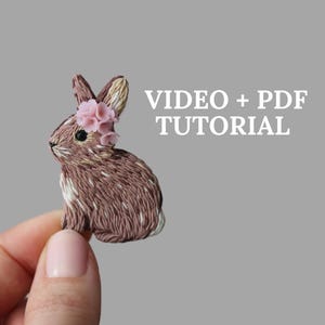 Könnte beinhalten: Ein braunes und weißes gesticktes Häschen mit rosa Blumenakzenten. Das Häschen wird von einer Hand gehalten. Der Text "VIDEO + PDF TUTORIAL" wird über dem Häschen angezeigt.