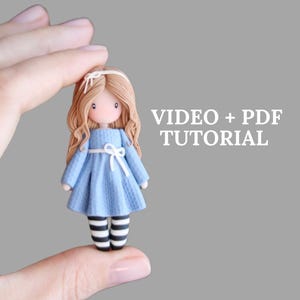 Puede incluir: Una muñeca hecha a mano con cabello castaño largo y ondulado, vestida con un vestido azul claro con un lazo blanco y medias a rayas blancas y negras. La muñeca está sostenida en una mano. El texto a la derecha dice "VIDEO + PDF TUTORIAL".