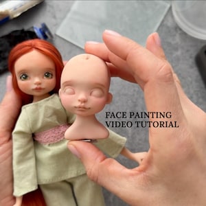 Può includere: Una bambola con capelli rossi e occhi verdi, che indossa un completo verde con una cintura rosa, è tenuta accanto a una testa di bambola. Il testo "FACE PAINTING VIDEO TUTORIAL" è visibile. I materiali per la pittura sono sullo sfondo.