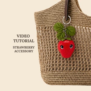 May include: A woven tan straw tote bag with a red strawberry keychain attached to the handle. The strawberry has a smiling face, large black eyes, and green fabric leaves. Text on the left reads: VIDEO TUTORIAL STRAWBERRY ACCESSORY.