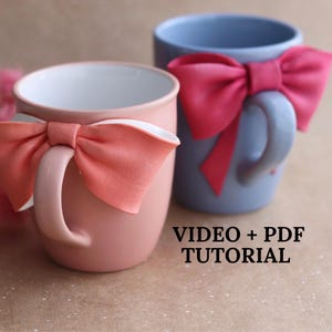 Könnte beinhalten: Zwei Keramikbecher, einer rosa und einer blau, jeweils mit einer großen Schleife verziert. Der rosafarbene Becher hat eine pfirsichfarbene Schleife, während der blaue Becher eine leuchtend rosa Schleife hat. Der Text "VIDEO + PDF TUTORIAL" ist sichtbar.