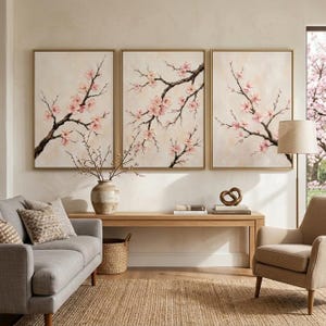 May include: Three framed wall art pieces featuring branches with pink blossoms against a neutral background. The artwork is displayed above a wooden console table, with a gray sofa, beige armchair, and a woven rug in the foreground. A vase with branches is on the table.