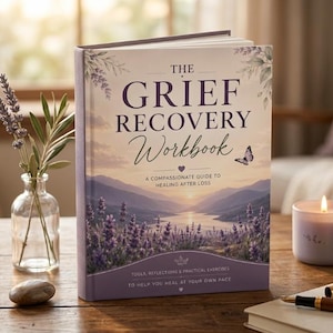 May include: A hardback book titled The Grief Recovery Workbook: A Compassionate Guide to Healing After Loss. The cover features a landscape of lavender fields and a lake at sunset, with a small butterfly, text, and purple accents.