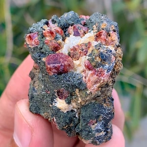 May include: A rough, dark gray rock with embedded red crystals. The crystals are clustered together and have a shiny, translucent appearance.