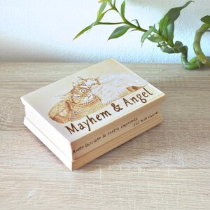 Può includere: Una scatola rettangolare in legno con finitura a venature chiare. Il coperchio presenta un'illustrazione stampata di due gatti, uno tigrato arancione e uno bianco, con il testo Mayhem & Angel e una scritta in italiano sul lato.
