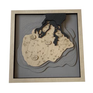 May include: A wooden wall art piece depicting a map of an island with a black outline. The island is surrounded by water with layered waves, and the island itself is filled with small squares representing buildings. The piece is framed in a light wood frame.