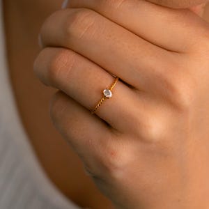 Puede incluir: Un anillo de oro con una pequeña gema transparente de forma ovalada. La banda del anillo tiene un diseño de cuerda trenzada. El anillo está en el dedo de una persona, con la mano descansando cerca de la cara.