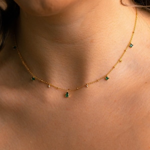 Puede incluir: Un delicado collar dorado con piedras preciosas verdes esmeralda y transparentes. La fina cadena tiene pequeñas cuentas doradas espaciadas uniformemente. Las piedras preciosas son una mezcla de formas cuadradas y de lágrima, añadiendo un toque de elegancia.