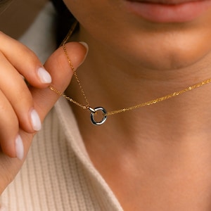 Puede incluir: Un delicado collar de oro con un pequeño colgante bicolor. El colgante es un círculo con la parte superior dorada e inferior plateada. El collar está sostenido por una mano, mostrando el diseño y los detalles de la joya.