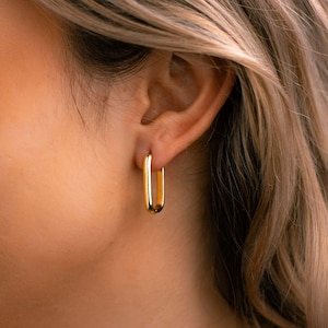 Könnte beinhalten: Nahaufnahme eines goldfarbenen, rechteckigen Creolen-Ohrrings. Der Ohrring hat ein schlichtes, poliertes Design mit glatter Oberfläche. Der Ohrring wird am Ohr einer Person mit blondem Haar getragen.