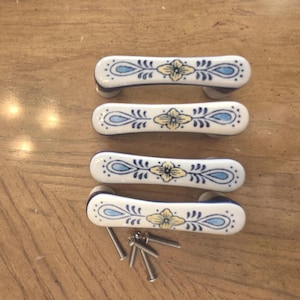 Puede incluir: Cuatro tiradores de cajón de cerámica blanca con forma alargada y curva. Cada tirador presenta un motivo floral pintado a mano en tonos azules y amarillos, acentuado por líneas decorativas de color azul oscuro. Incluye cuatro tornillos.