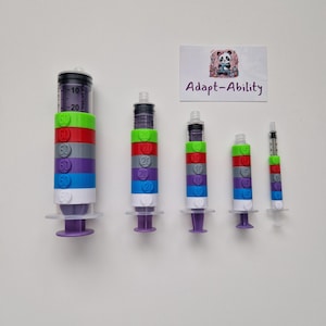 May include: Five syringes with colourful, stacked rings. Each syringe has a clear barrel with graduated markings and a plunger. The rings are in various colours, including green, red, grey, purple, and white. The text "Adapt-Ability" is on a small card.