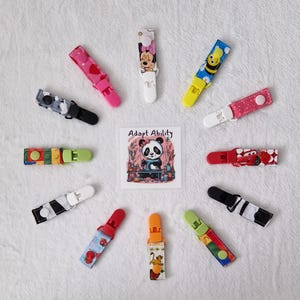 May include: A selection of baby pacifier clips in a variety of colours and patterns, including cartoon characters and a panda bear illustration. The clips are arranged in a circle on a white background.