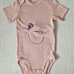 May include: Pale pink baby bodysuit with short sleeves and a zip pocket. A purple medical device is attached to the pocket. The bodysuit has popper fastenings at the bottom and a tag attached at the top.