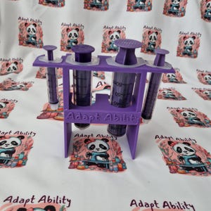 May include: A purple syringe holder with three syringes. The holder has the text "Adapt Ability" on the front. The syringes are clear with black measurements. The background is a white fabric with panda bear illustrations.