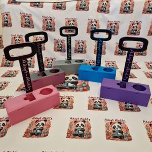 May include: Four rectangular plastic stands in pink, grey, blue, and purple, each featuring a vertical handle with the text Adapt-Ability printed on it. The stands have a circular cutout and a rectangular slot on the top surface.