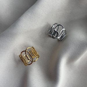 Könnte beinhalten: Zwei breite Ringe, einer in poliertem Goldton und einer in poliertem Silberton. Jeder Ring verfügt über ein durchbrochenes Design aus mehreren miteinander verbundenen ovalen Schlaufen, die sich um den Finger legen und ein durchgehendes, luftiges und strukturiertes Metallmuster bilden.