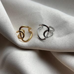 Puede incluir: Dos anillos abstractos, uno dorado y otro plateado, descansan sobre una tela neutra. Los anillos presentan un diseño continuo en bucle con un elemento en forma de nudo. El anillo dorado está a la izquierda y el plateado a la derecha.