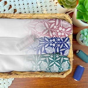 Peut inclure: Trois panneaux de tissu pliés dans un panier en osier, chacun présentant un dégradé blanc vers une couleur avec un motif botanique de grenades et de fleurs blanches. Les couleurs passent du rose au bleu puis au sarcelle, avec trois bobines.