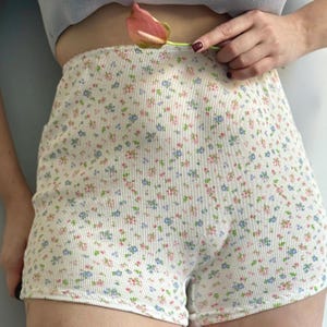 Puede incluir: Pantalones cortos de cintura alta con estampado floral. Los pantalones cortos son blancos con una textura acanalada y están cubiertos de pequeñas flores azules, rosas y verdes. Los pantalones cortos están hechos de un material suave y cómodo.