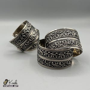 Può includere: Bracciali a polsino argentati con intricati motivi floreali e geometrici. L'immagine mostra tre bracciali larghi, uno inclinato e gli altri impilati. I bracciali hanno un'estetica vintage.