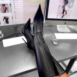 May include: Black leather knee-high boots with pointed toes and stiletto heels. The boots feature a side zip. The image is set against a blurred background.