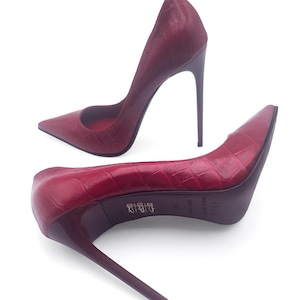 Peut inclure: Une paire d'escarpins rouges à talons hauts avec un bout pointu et un talon aiguille. Les chaussures présentent un motif texturé en forme de peau de crocodile. Les talons mesurent environ 10 cm de haut.