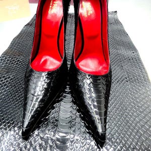 Può includere: Un paio di scarpe col tacco nere con una finitura lucida e strutturata. L'interno è di un rosso vibrante e le scarpe hanno la punta a punta. I tacchi sono alti e sottili e le scarpe sono esposte su una superficie strutturata.
