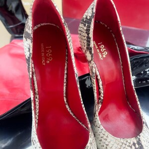 May include: Pair of high-heeled pumps with a snakeskin pattern in beige and brown. The interior is red, and the shoes are marked "1969 Made in Italy". The heels are pointed and the shoes are displayed on a reflective surface.