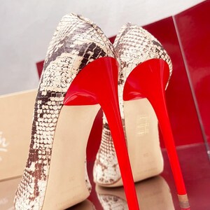 May include: A pair of high-heeled shoes with a snakeskin pattern in shades of brown and cream. The shoes feature bright red soles and stiletto heels, creating a striking contrast. The heels are approximately 4 inches tall.