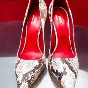 May include: A pair of high-heeled pumps with a snakeskin pattern in shades of brown, black, and white. The interior is a vibrant red, and the shoes have pointed toes. The text "1969. MADE IN ITALY" is visible.