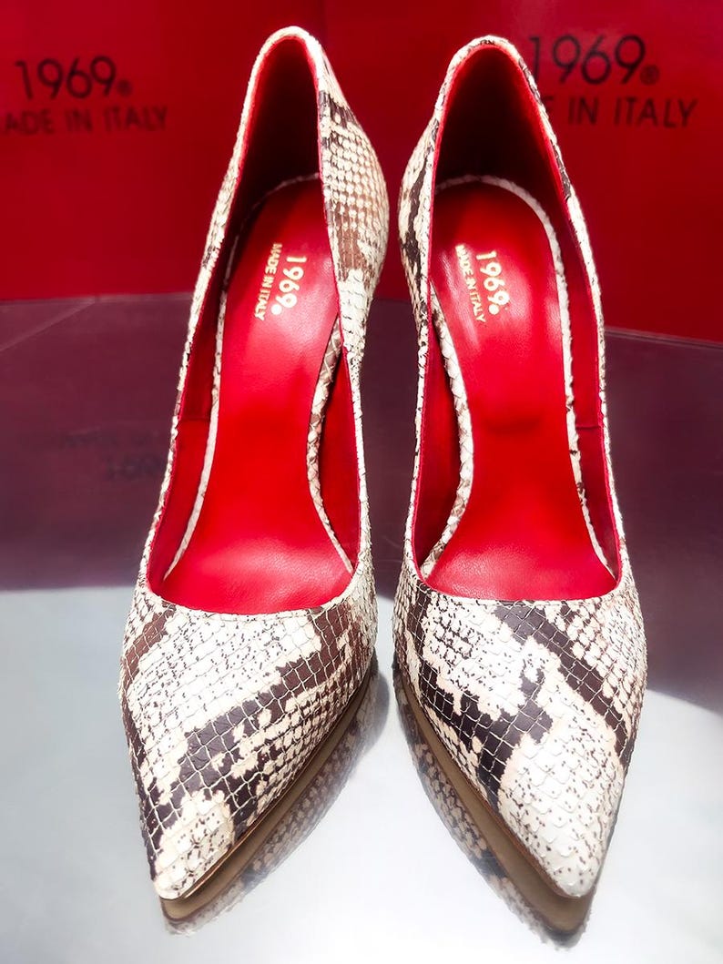 May include: A pair of high-heeled pumps with a snakeskin pattern in shades of brown, black, and white. The interior is a vibrant red, and the shoes have pointed toes. The text "1969. MADE IN ITALY" is visible.