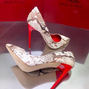 May include: A pair of high-heeled shoes with a snakeskin pattern in white, brown, and tan. The shoes have bright red heels and soles. The text "1969. MADE IN ITALY" is visible in the background.