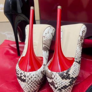 May include: A pair of high-heeled shoes with a snakeskin pattern and bright red soles and heels. The shoes are set against a red background, with a black boot visible in the background.