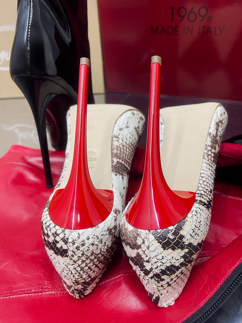 May include: A pair of high-heeled shoes with a snakeskin pattern and bright red soles and heels. The shoes are set against a red background, with a black boot visible in the background.