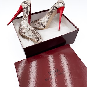 May include: A pair of high-heeled pumps with a snakeskin pattern in a burgundy box. The shoes have red heels and soles. The box is burgundy with the text "MADE IN ITALY".