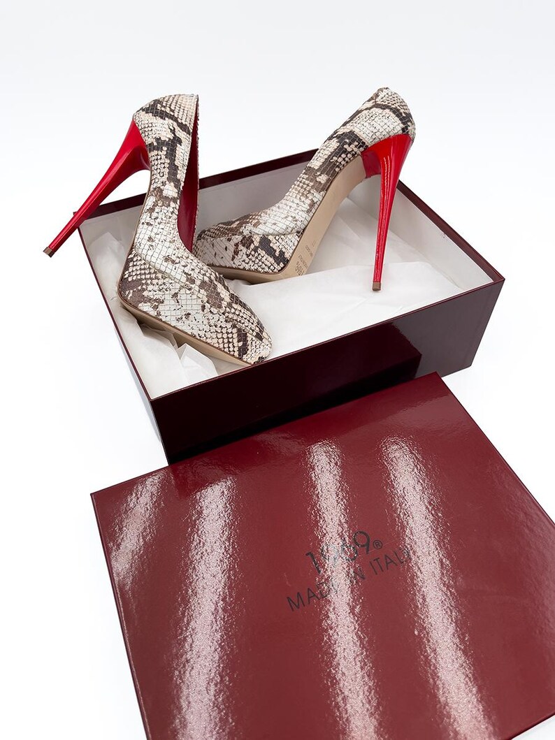 May include: A pair of high-heeled pumps with a snakeskin pattern in a burgundy box. The shoes have red heels and soles. The box is burgundy with the text "MADE IN ITALY".