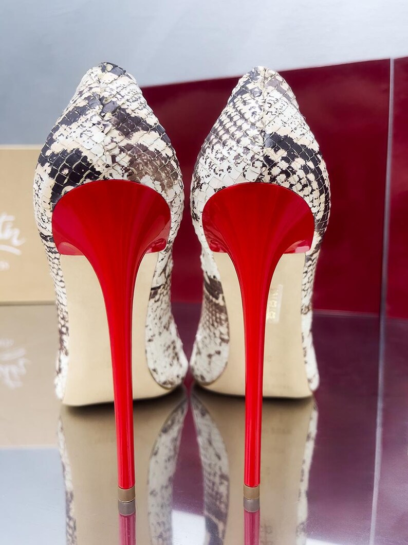 May include: A pair of high-heeled shoes with a snakeskin pattern in black, white, and brown. The shoes have bright red stiletto heels and a matching red interior. The shoes are displayed on a reflective surface.