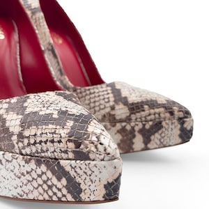 May include: Close-up of two high-heeled pumps with a snakeskin pattern in shades of brown, beige, and black. The interior of the shoes is a vibrant red. The shoes have a platform sole.