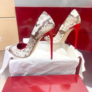 May include: A pair of high-heeled shoes with a snakeskin pattern in shades of brown and white. The shoes have bright red heels and are displayed in a red box with the text "1969 MADE IN ITALY".
