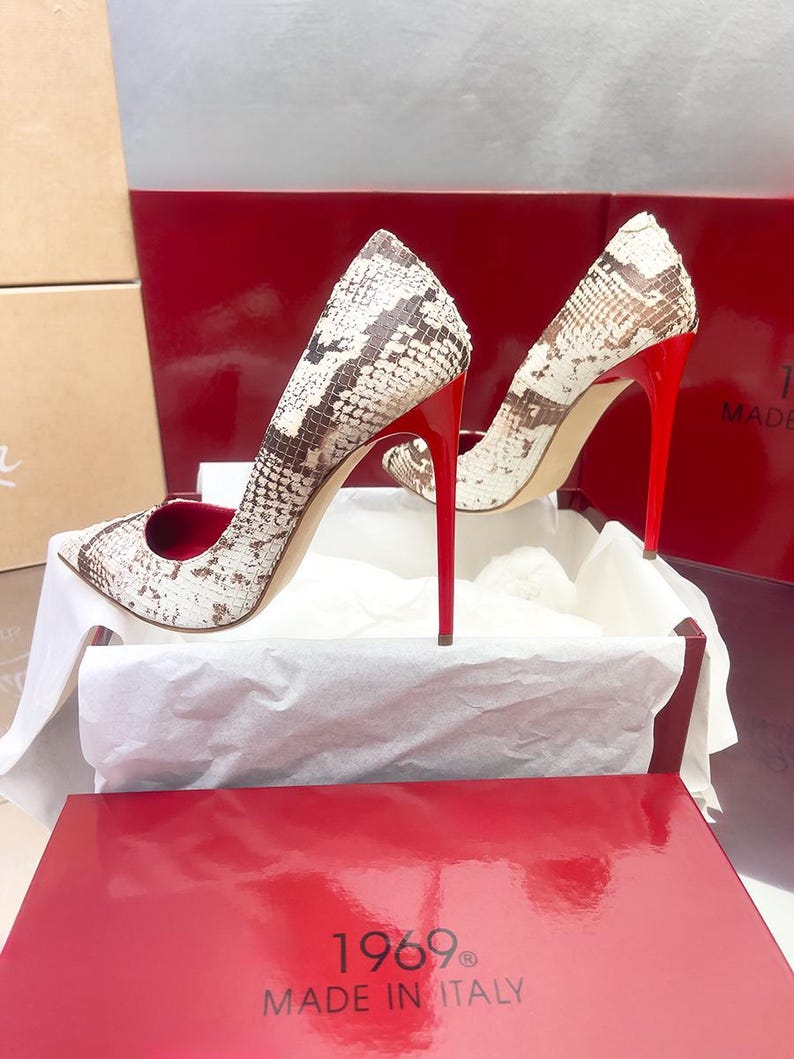 May include: A pair of high-heeled shoes with a snakeskin pattern in shades of brown and white. The shoes have bright red heels and are displayed in a red box with the text "1969 MADE IN ITALY".