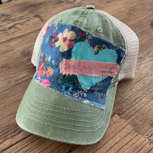May include: A green and beige trucker hat with a floral fabric patch and the word "courage" written on a pink fabric patch. The hat has a mesh back.