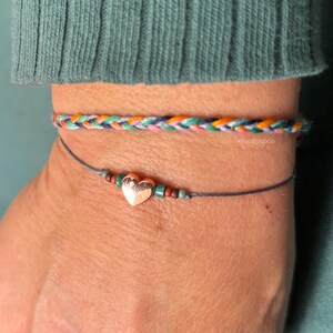 May include: Two bracelets: one is a multi-colored braided cord featuring pink, orange, green, and dark blue threads. The second is a thin grey string bracelet with a small metallic heart charm flanked by tiny green and brown seed beads.