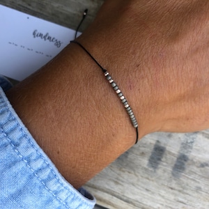 May include: A simple bracelet with a black cord and small gray and rose gold beads. The bracelet is tied with a slip knot.