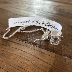 May include: A silver necklace with a small, round, silver and gold charm that says "great is thy faithfulness" on a white tag. The charm is a small box with a heart on the front.