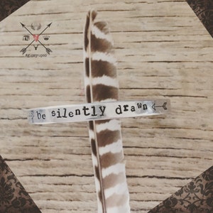 May include: A silver metal bracelet stamped with the words "be silently drawn" and an arrow design. The bracelet rests on a brown and white feather, set against a wooden background. The image also includes a logo with text.